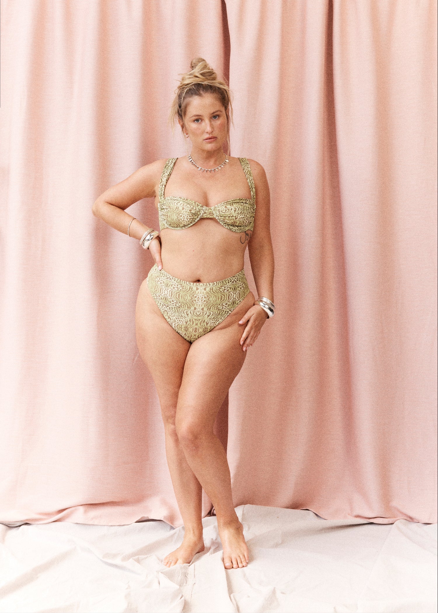 A woman models a two-piece bikini set with a textured, metallic gold and olive green pattern, standing against a draped pink fabric backdrop.