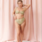 A woman modeling a two-piece bikini set with a textured, metallic gold and olive green pattern, standing against a draped pink fabric backdrop.