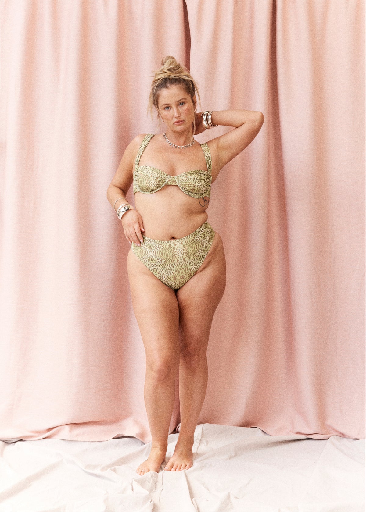 A woman modeling a two-piece bikini set with a textured, metallic gold and olive green pattern, standing against a draped pink fabric backdrop.