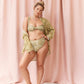 A woman models an olive-green sequined bikini set with a matching open-front cover-up against a draped pink fabric backdrop.