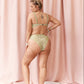 A woman stands barefoot in front of pink draped curtains, wearing a two-piece bikini with a green and gold floral pattern. She is shown from the back, posing with her hands on her hips.