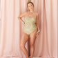A woman models an olive-green one-piece swimsuit with spaghetti straps, standing barefoot in front of a draped pink fabric backdrop.