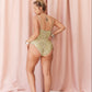 A woman stands barefoot in front of pink draped curtains, photographed from the back while wearing a one-piece swimsuit with a textured green and beige pattern.