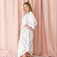 A woman modeling a white kaftan dress with colorful floral patterns, standing in front of a dusty pink curtain backdrop.