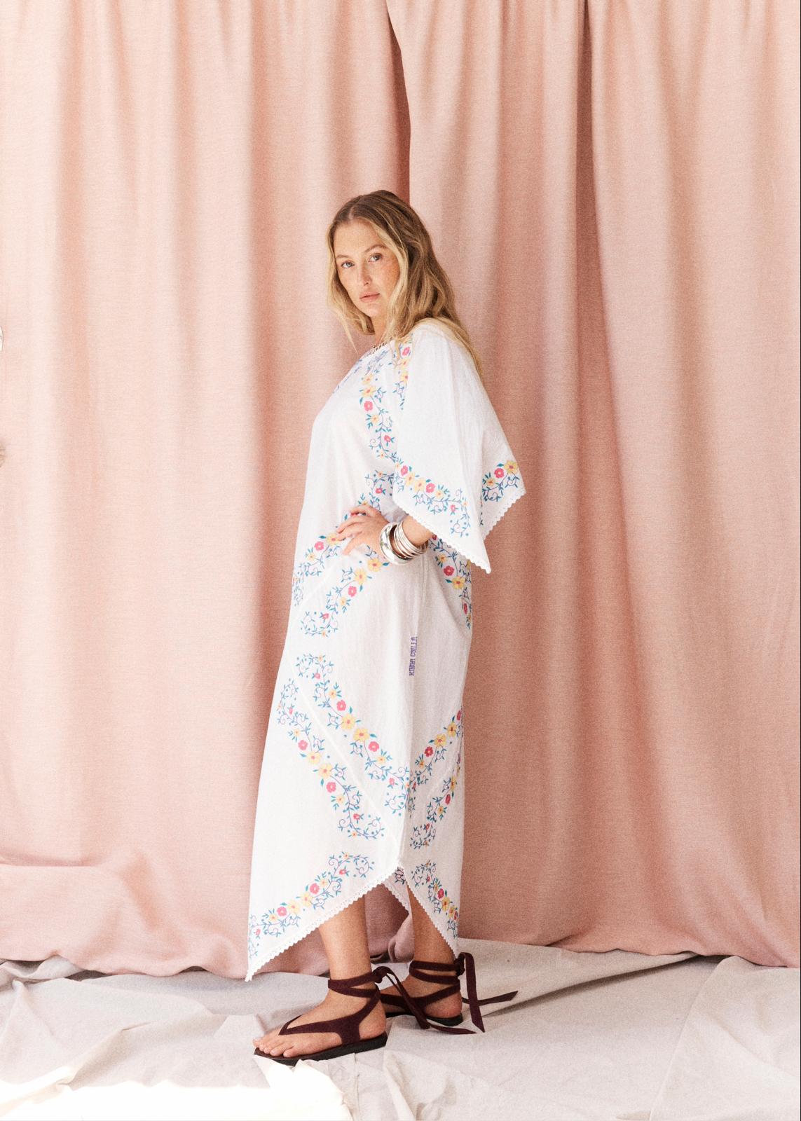 A woman modeling a white kaftan dress with colorful floral patterns, standing in front of a dusty pink curtain backdrop.