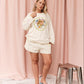 A woman modeling a cream-colored lounge set consisting of a long-sleeve sweatshirt and matching shorts, featuring a colorful graphic print on the sweatshirt. She is standing in front of a pink curtain backdrop with a small metallic stool to her side.