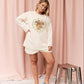 A woman models a cream-colored lounge set consisting of a long-sleeve sweatshirt and matching shorts, featuring a colorful graphic of a dolphin and other marine wildlife, with the word 'KINGA' on the front. She is standing in front of a pink curtain backdrop with a small metallic stool to her side.