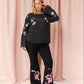 A woman modeling a black long-sleeve top and black flared pants set with colorful embroidered details, standing against a pink draped fabric backdrop.