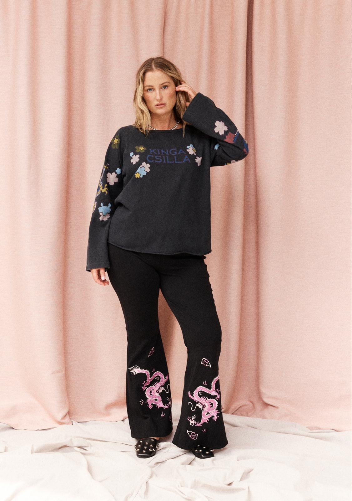 A woman modeling a black long-sleeve top and black flared pants set with colorful embroidered details, standing against a pink draped fabric backdrop.