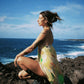 Woman in a colorful dress sitting on rocks by the ocean with a blue sky.