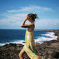 Woman in a yellow slip dress and black boots standing on rocky coastal landscape with ocean and blue sky.