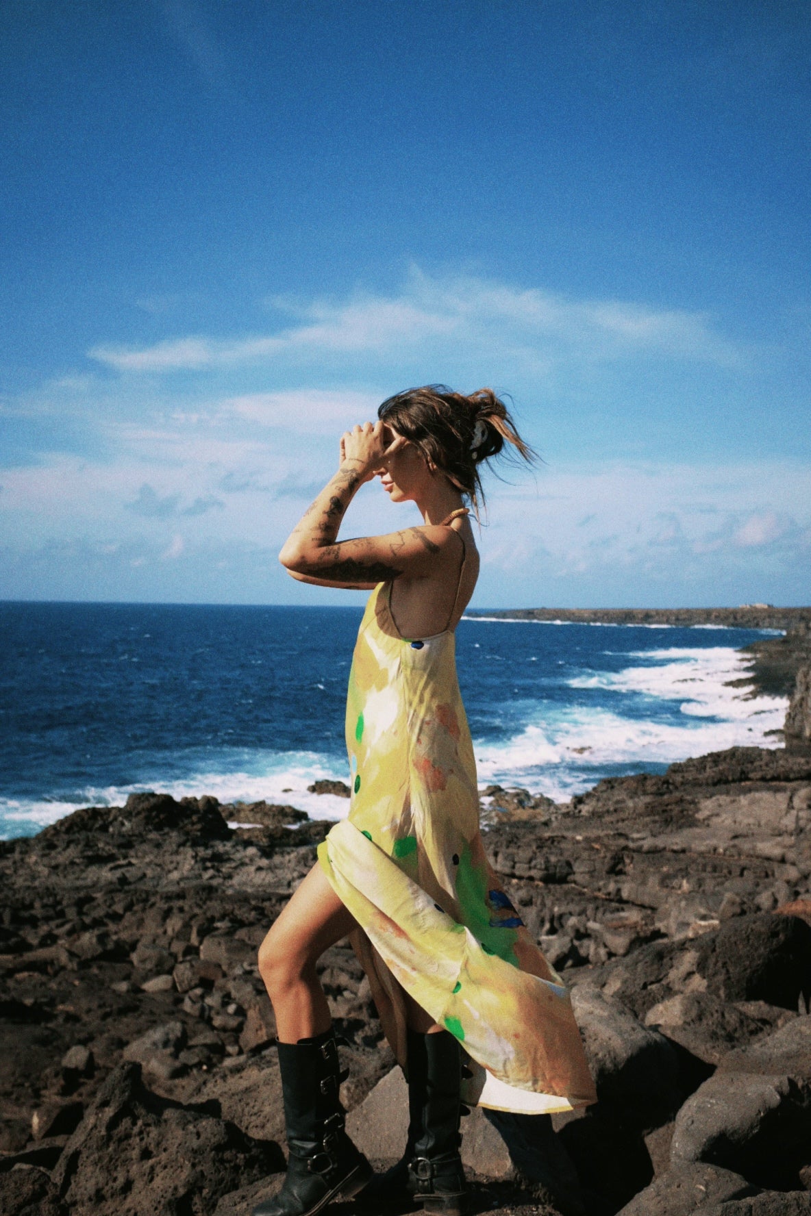 Woman in a yellow slip dress and black boots standing on rocky coastal landscape with ocean and blue sky.