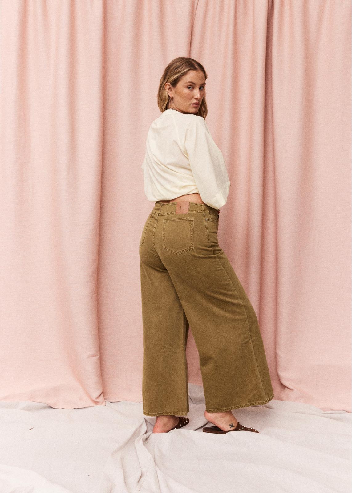 A woman stands in profile wearing wide-leg pants in an olive-brown denim fabric, paired with a white cropped blouse and flat sandals, set against a draped pink fabric backdrop.