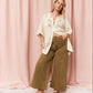 A woman stands in front of a pink draped fabric backdrop wearing a cream embroidered short-sleeve top and olive wide-leg pants, with brown flat shoes.