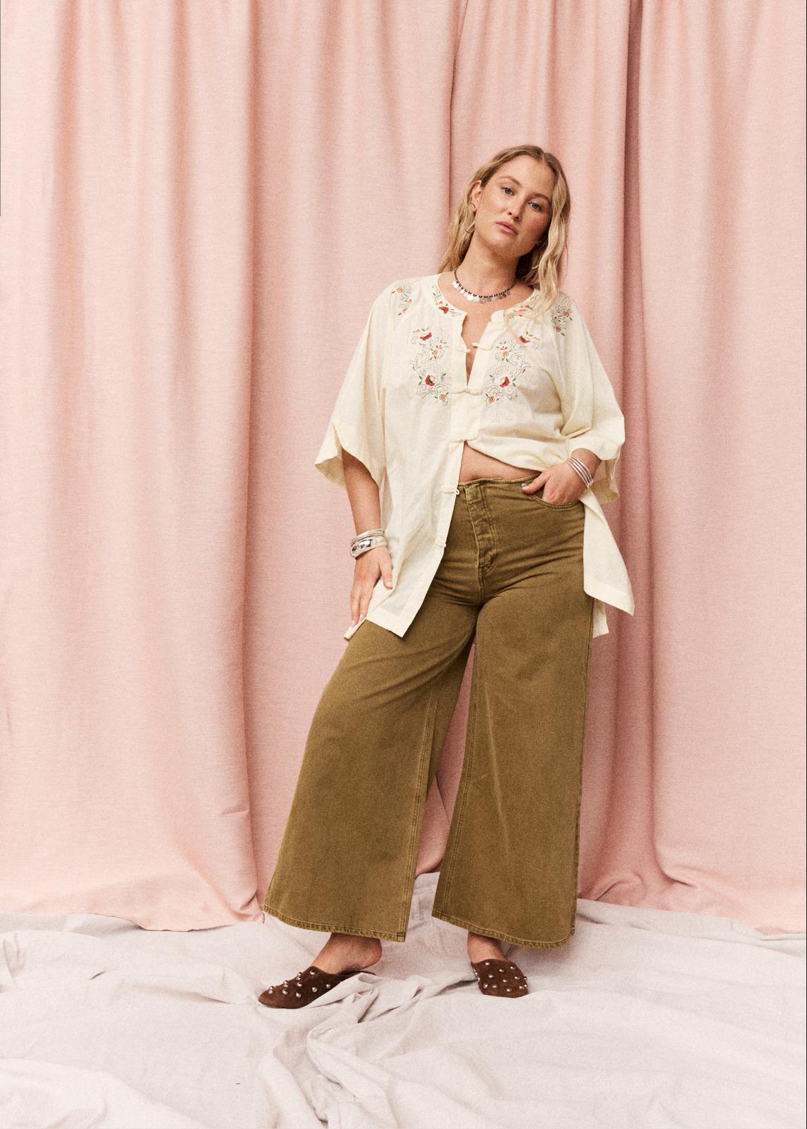 A woman stands in front of a pink draped fabric backdrop wearing a cream embroidered short-sleeve top and olive wide-leg pants, with brown flat shoes.