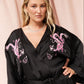 A woman wearing a black silk-like wrap robe with pink embroidered dragon motifs on the shoulders, posed against a soft pink draped backdrop.