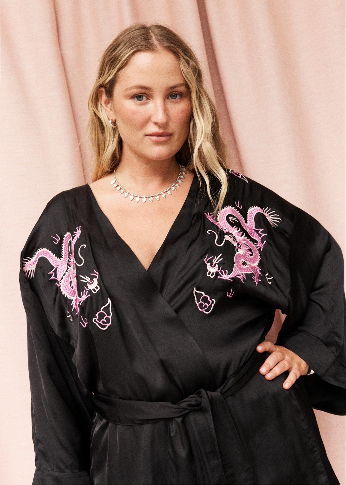 A woman wearing a black silk-like wrap robe with pink embroidered dragon motifs on the shoulders, posed against a soft pink draped backdrop.