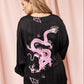 A woman shown from the back wearing a black long-sleeve top with a large pink dragon graphic and small cloud motifs, posed against a pink curtain backdrop.