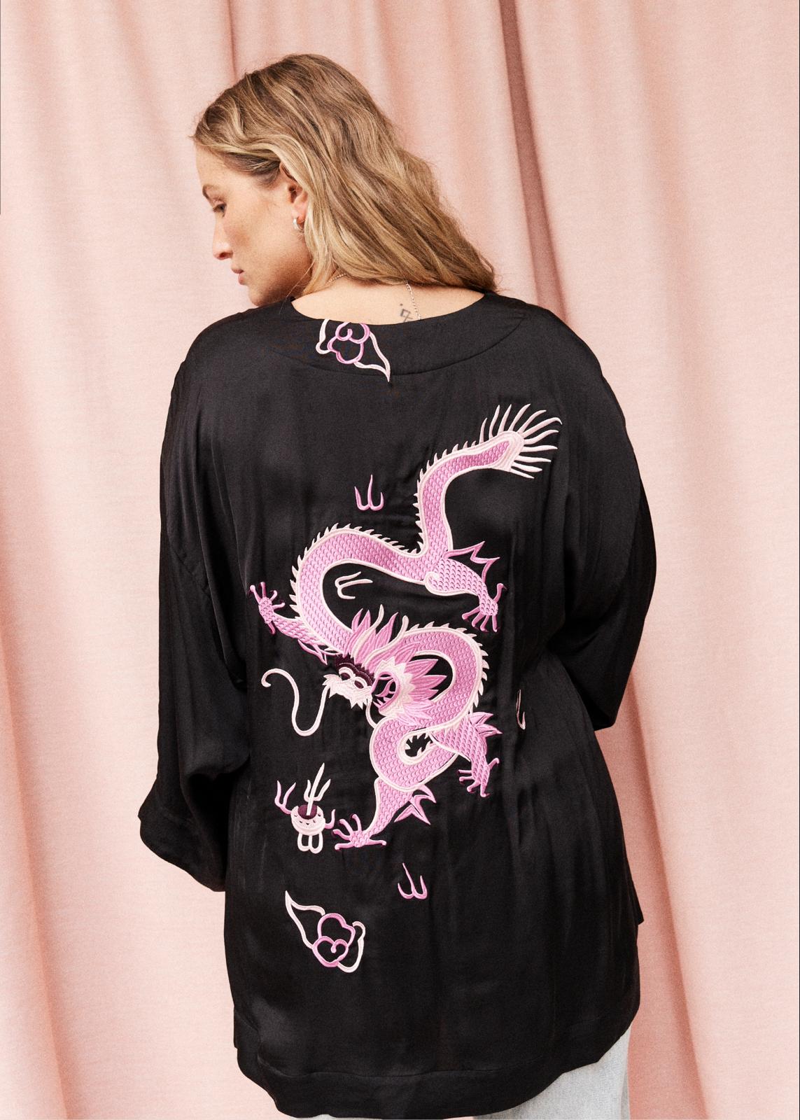 A woman shown from the back wearing a black long-sleeve top with a large pink dragon graphic and small cloud motifs, posed against a pink curtain backdrop.
