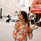 Model wearing Kinga Csilla Farfalla Olive Dress, a long-sleeve orange maxi with butterfly and floral print, holding baguettes on a Paris street with the Eiffel Tower blurred in the background.