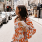 Model wearing the Kinga Csilla Farfalla Olive Dress, a flowing orange maxi with butterfly and floral motifs, carrying baguettes on a sunlit Paris street with the Eiffel Tower behind.
