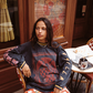 Model wearing Kinga Csilla Love Potion Roses Tee in stonewashed black with graphic rose print, styled with Farfalla Jessi Pant in orange butterfly cloud print, sitting at a Parisian café