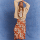 Model wearing the Kinga Csilla Shell OG Jumper, a cream oversized cotton sweater with hand-drawn shell and sun doodle graphic, styled with bold orange floral Farfalla Olive Dress and brown sandals. Relaxed bohemian streetwear look against a textured blue backdrop.