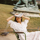 Model in the Kinga Csilla Tottori Lace Harlow Maxi Dress in white cotton lace, sitting on a Parisian garden chair holding a newspaper with a bronze statue in the background.