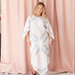 A woman modeling a white kaftan dress with colorful floral embroidery, standing in front of a pink curtain backdrop.