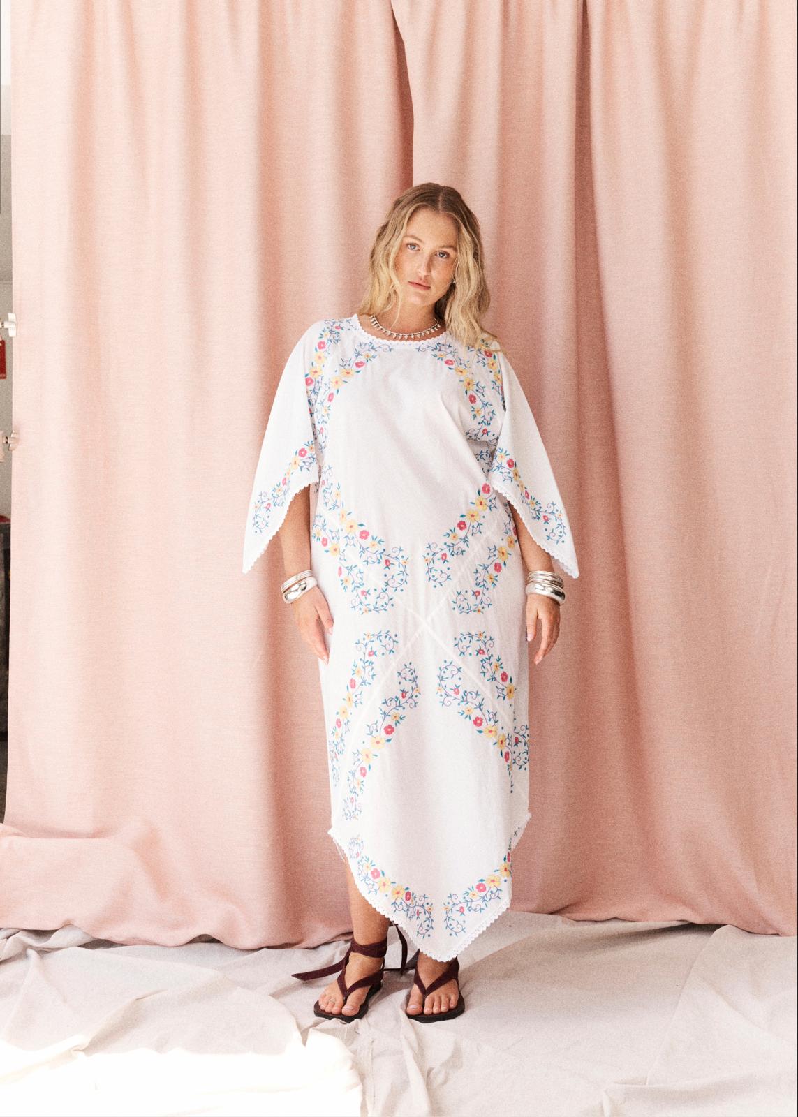 A woman modeling a white kaftan dress with colorful floral embroidery, standing in front of a pink curtain backdrop.